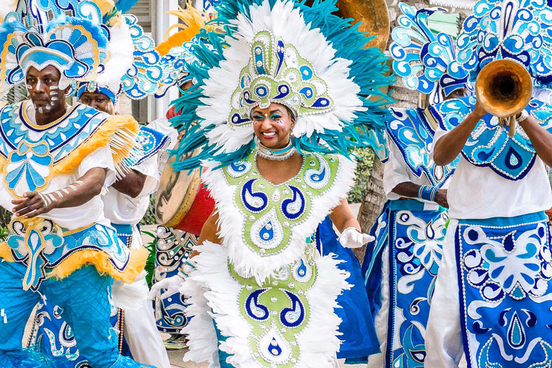 A colorful Junkanoo dancer wearing white feathers and green and blue regalia, with several Junkanoo musicians behind them, celebrating in the Bahamas.
