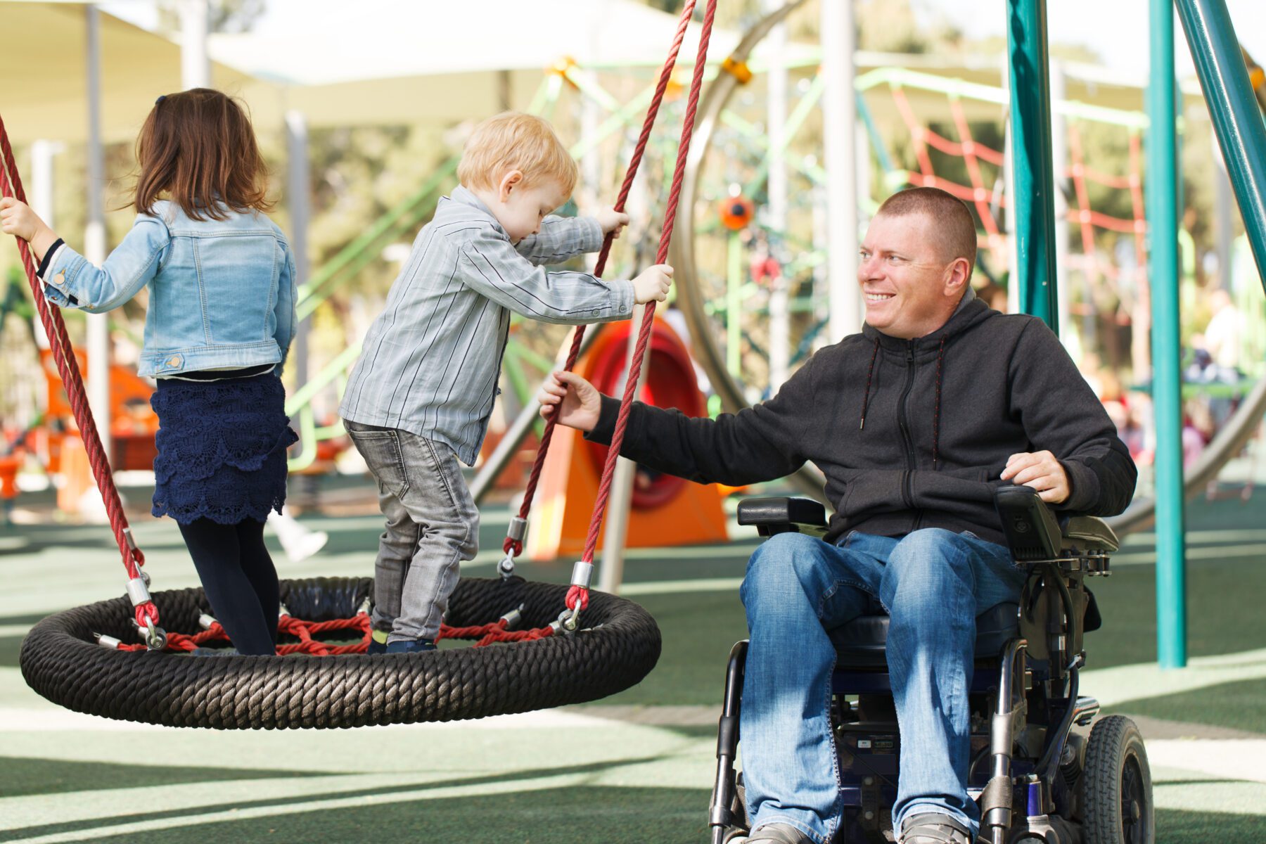 A man in a wheelchair pushes two children, a boy and a girl, on a tire swing at a playground.