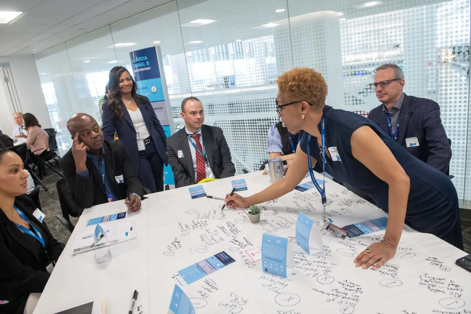 Black woman, pointing at documents on a table, while talking to a group of people in small conference room.