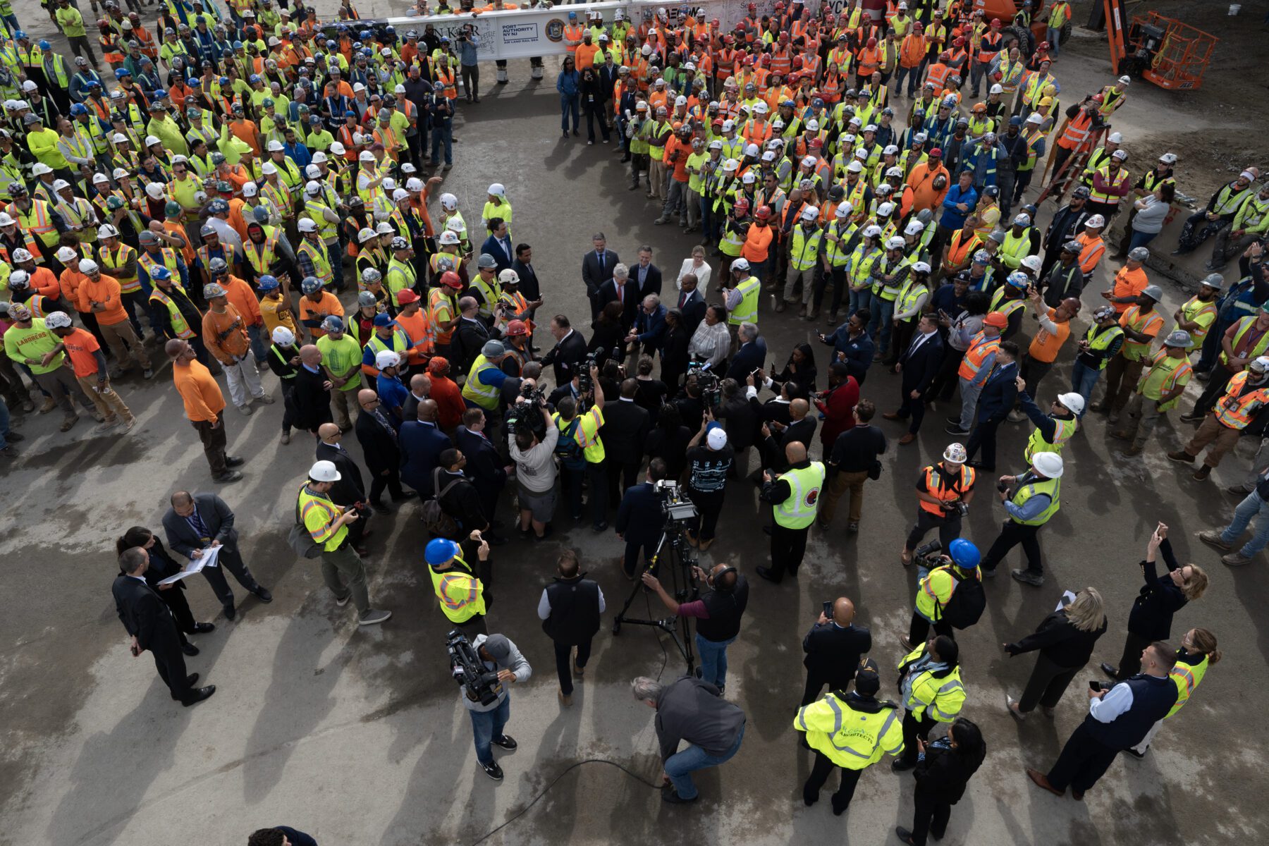 A large group of construction workers, all wearing safety vests and helmets, gather around a central group of people on a construction site. Various equipment and barriers are visible in the background.