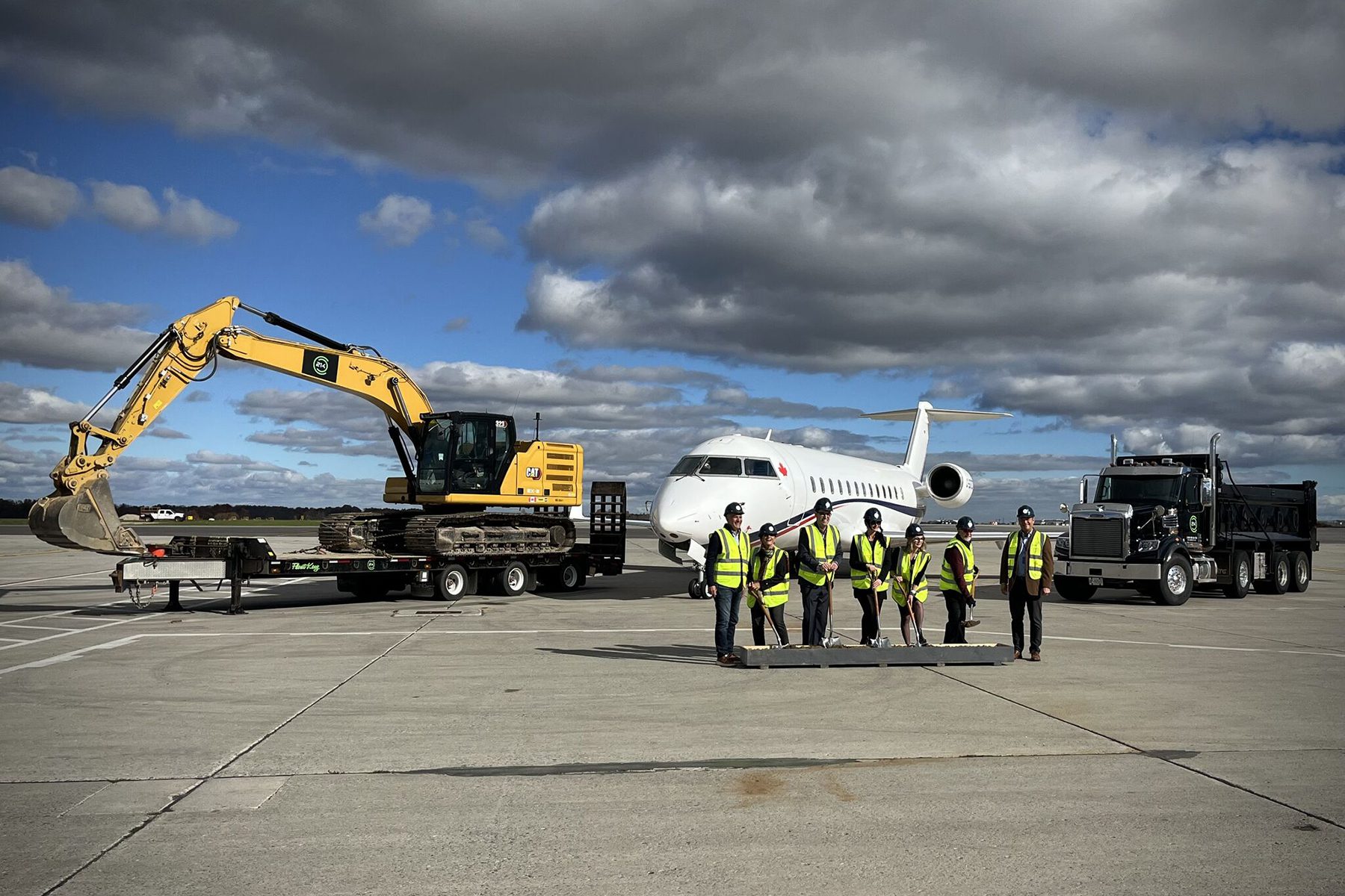 A group of people in safety vests stand on an airport tarmac between a large excavator on a truck, a parked private jet, and a dump truck under a cloudy sky.