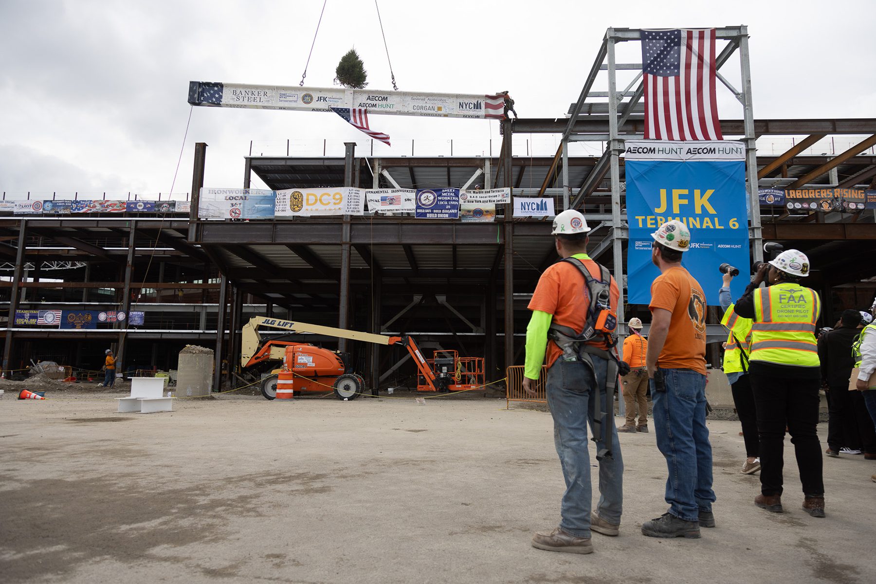 Construction workers at a building site observe a beam being hoisted. A tree is attached to the beam, and flags are displayed. Signs and banners are visible on the structure.