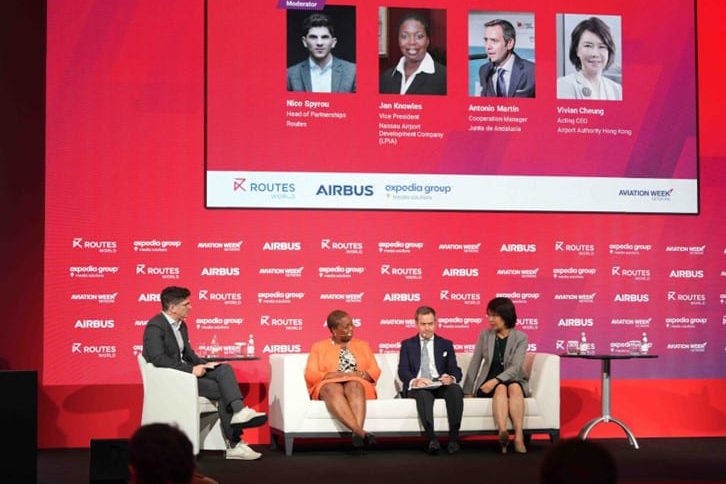 Four panelists and a moderator seated on stage discuss aviation topics at the World Routes 2023 event, with a red backdrop displaying logos and participant names.