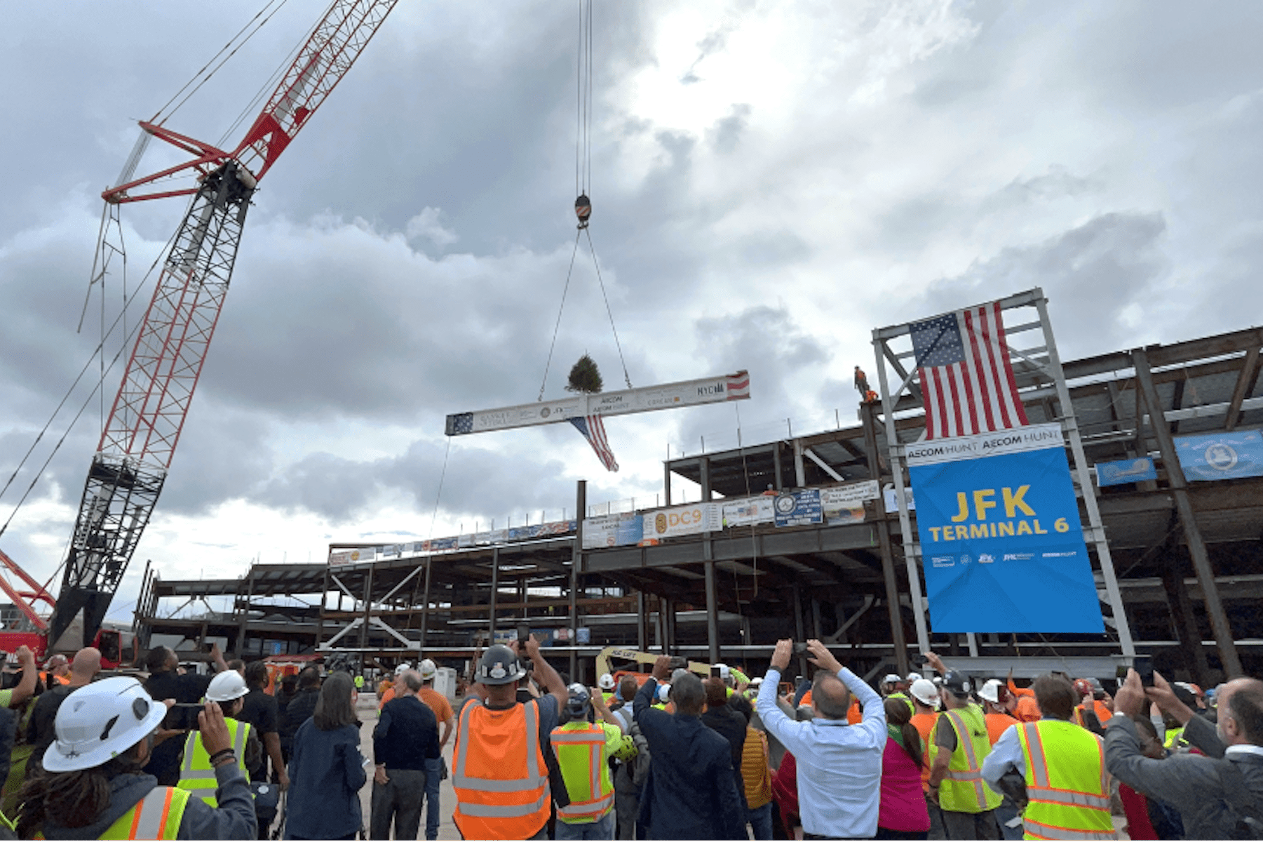 A construction crew at JFK Terminal 6 observes a steel beam being lifted by a crane during a topping-out ceremony, with people wearing hard hats and safety vests.