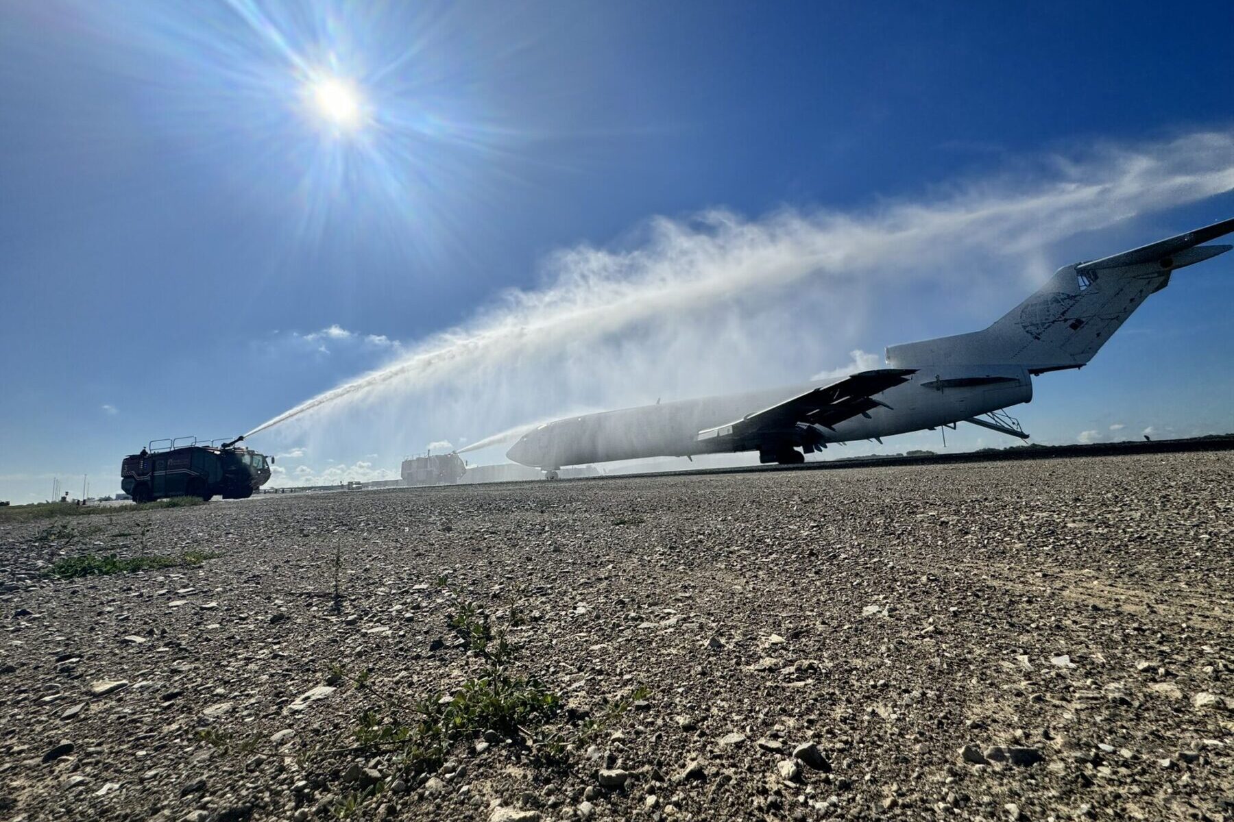 A fire truck sprays water on a large airplane on the ground under a bright sun.