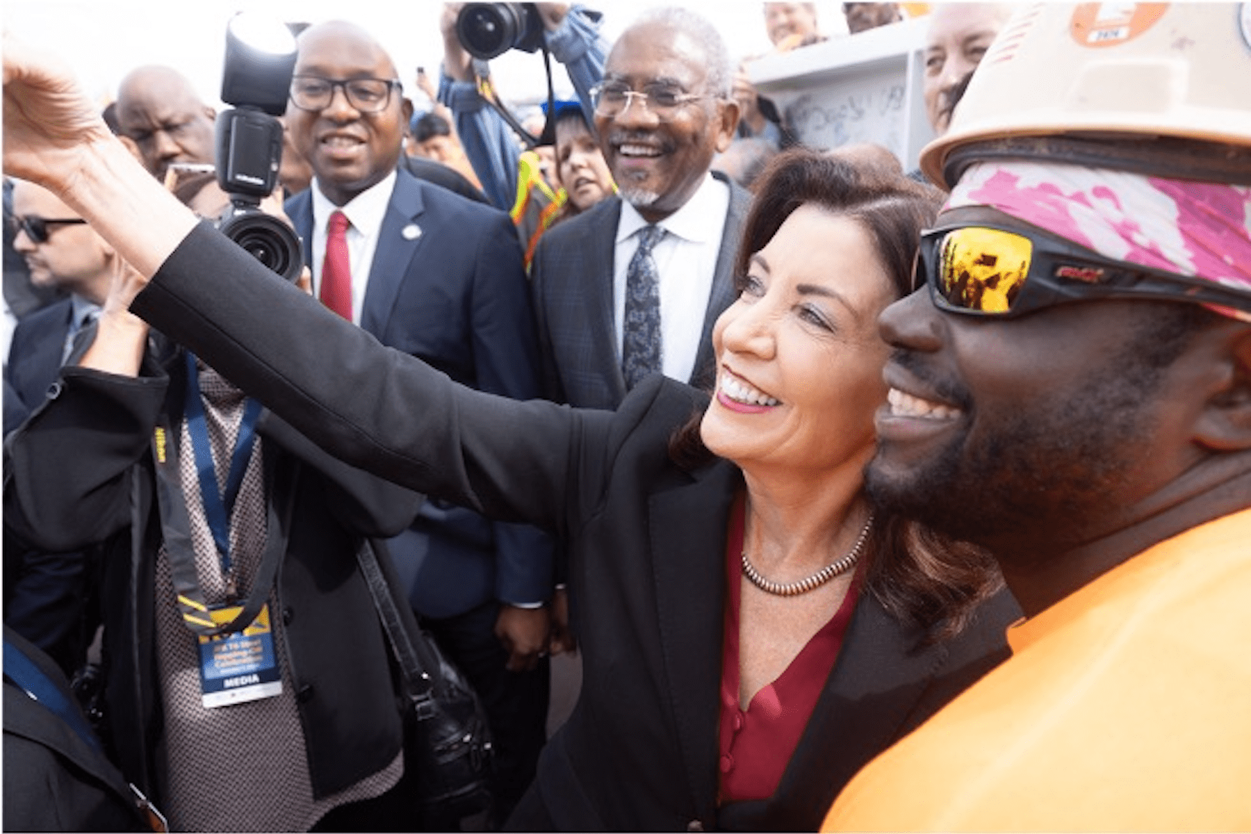 Gov Hochul takes a selfie with a construction worker wearing a helmet and sunglasses. Other people surround them, some smiling and looking at the camera.