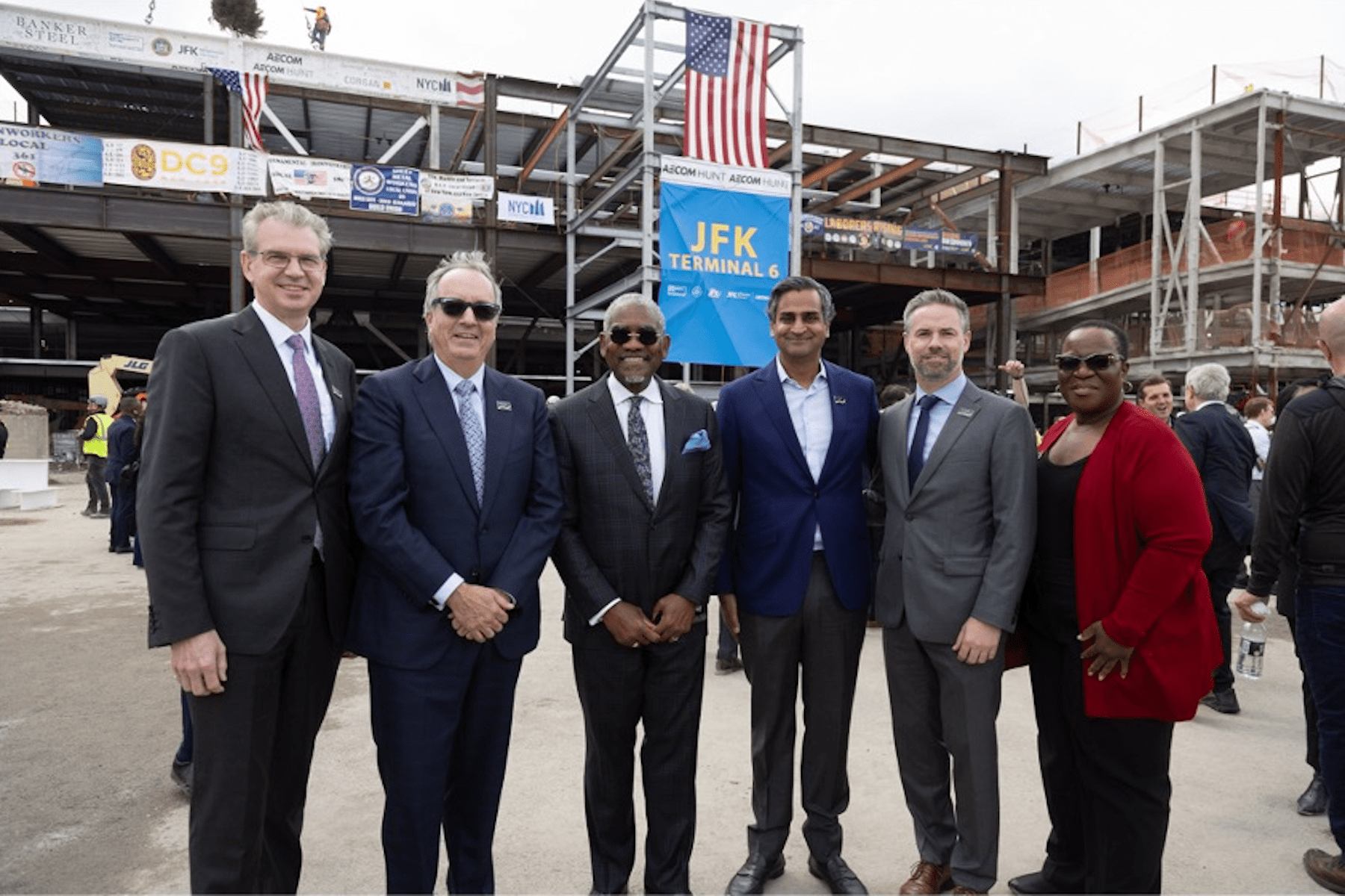 A group of Vantage Executives and Board members stand in front of a construction site for JFK Terminal 6, with steel framework and American flags visible.