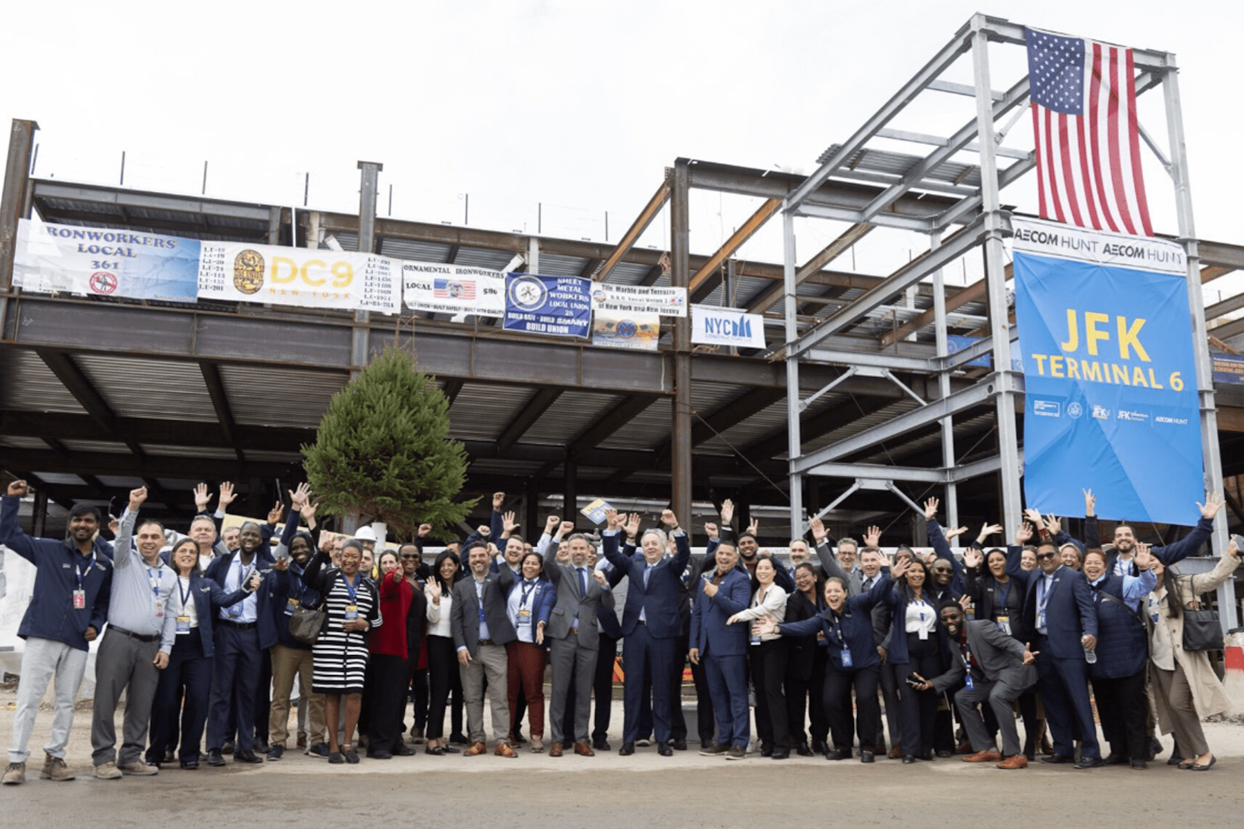 A large group Vantage Group and JFK Millennium Partners employees celebrate in front of an under-construction building labeled JFK Terminal 6, with an American flag displayed above.