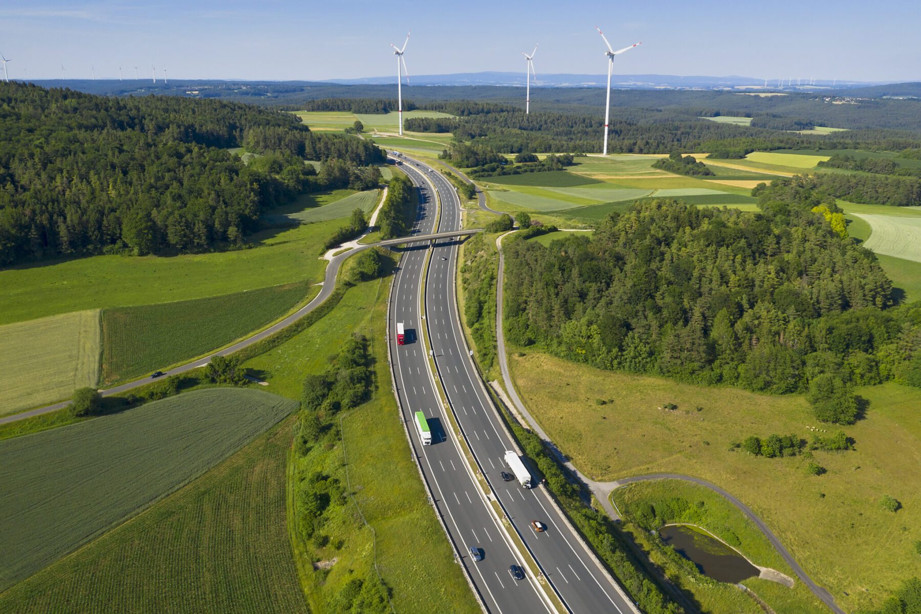 Aerial view of a highway with several vehicles, surrounded by green fields and forests. Wind turbines are visible in the background.