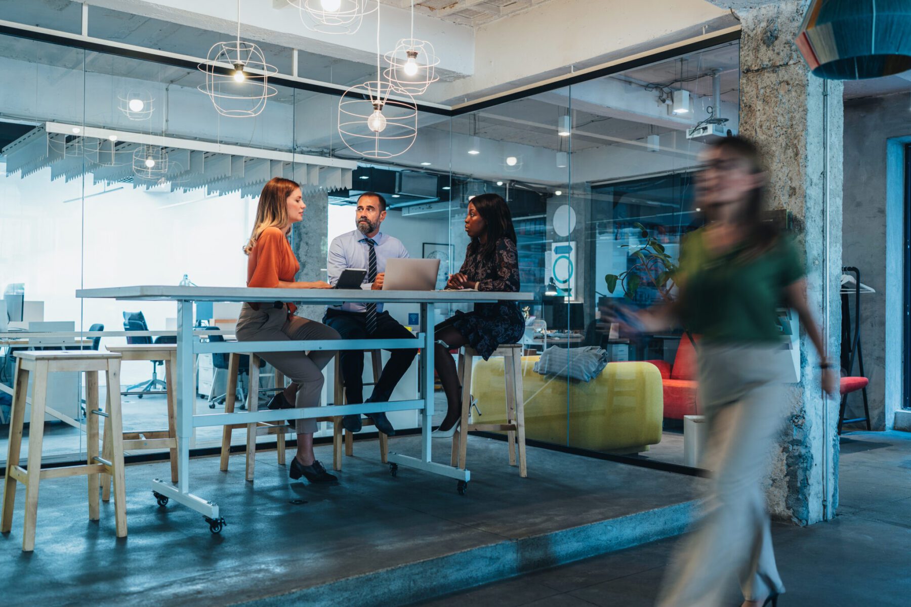 Three people sit at a high table in a modern office discussing with a laptop while another person walks by.