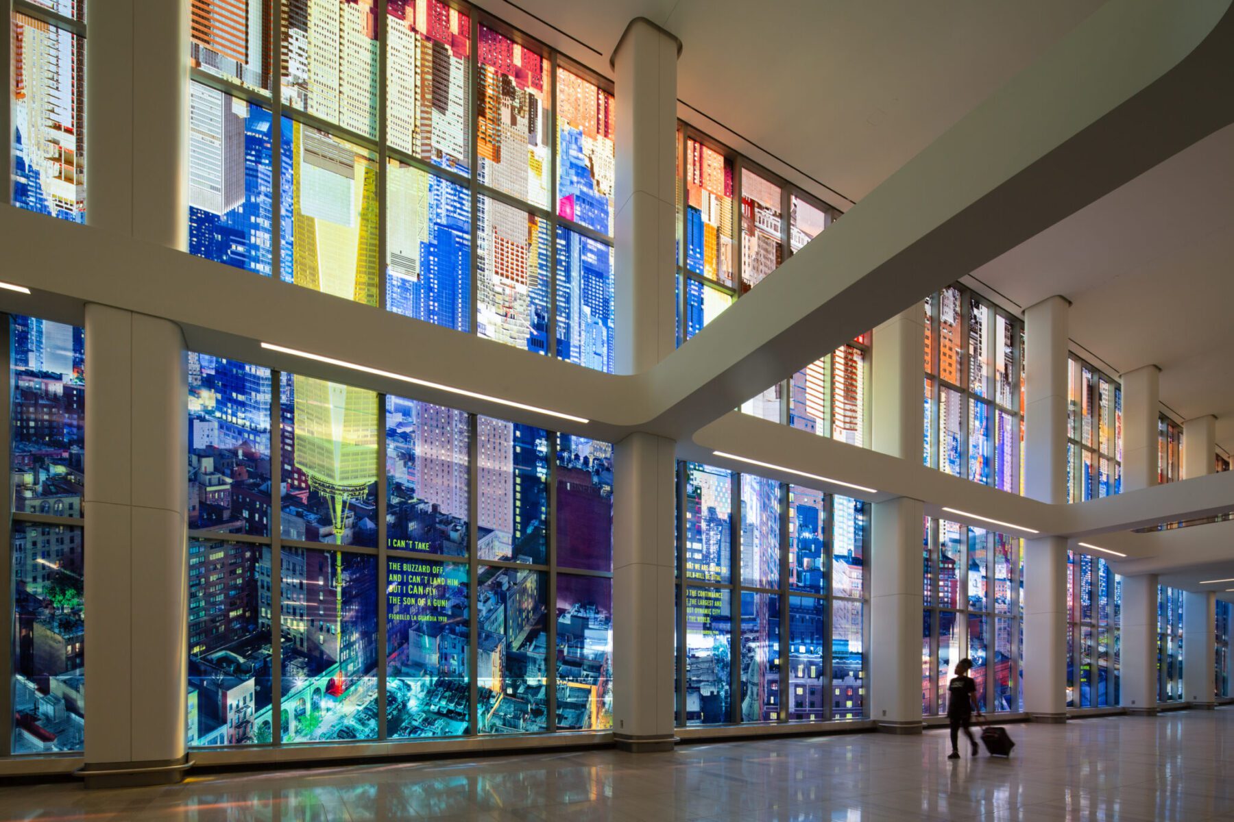 A person with a suitcase walks inside a large building with colorful, city-themed stained glass windows.