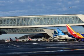 Several commercial airplanes parked at an airport terminal with a glass-clad bridge above. A plane is taking off in the background under a blue sky with clouds.