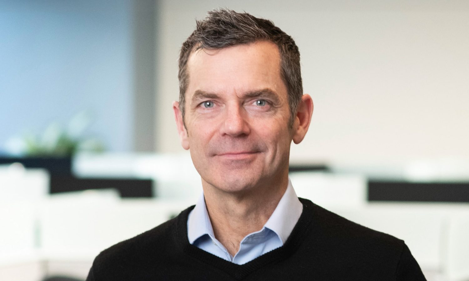 A person wearing a black sweater and blue shirt, standing indoors with blurry office background.