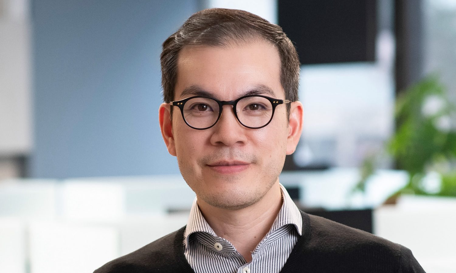 The image shows a person wearing glasses and a black sweater over a striped shirt, standing in an office setting.